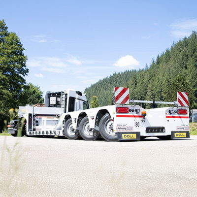 Weißer Sattelschlepper mit dreiachsigem Anhänger auf einer sonnigen Straße, umgeben von üppigen grünen Bäumen, unter einem klaren blauen Himmel.